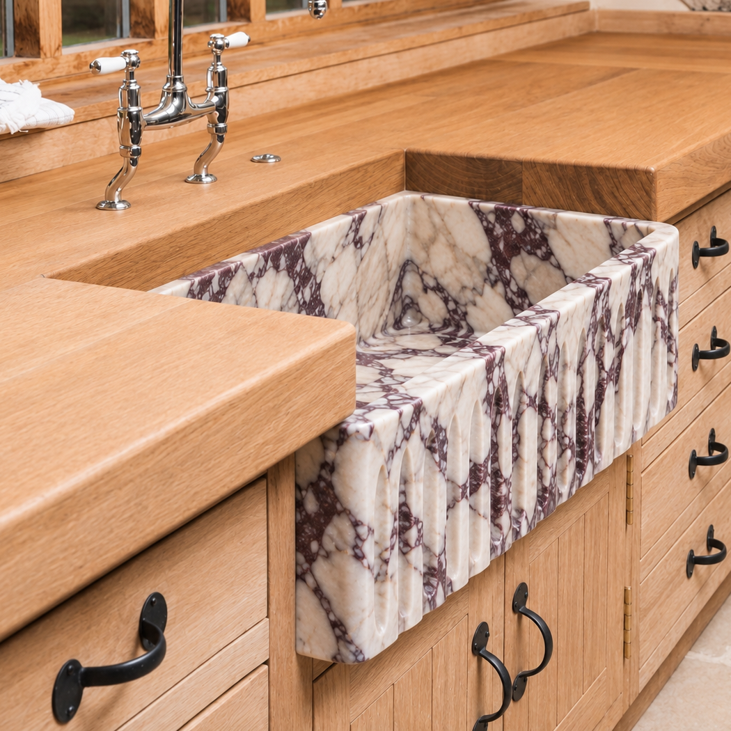 A close-up of a farmhouse-style kitchen sink with a white and dark gray veined marble basin set into a warm wood countertop and matching cabinets.